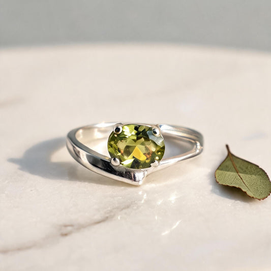 A sterling silver ring with a round-cut olive green gemstone, resting on a white marble surface next to a small green leaf.